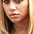 Static pixelated image of a person with light brown hair, fair skin, and dark eyes wearing a silver chain necklace against a blurred background.