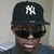 A person wearing a black cap with white NY logo, dark sunglasses, and gray shirt in four- view