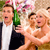 Two people in formal attire pose together; one holds a green bottle and pink flowers, with a blurred background.