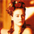 Pixelated image of a person with red hair styled in an updo, wearing a dark top against a warm-toned background.