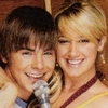 Two people pose closely together, one holding a silver microphone, both smiling against a brown background.