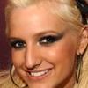 A close-up of a person with blonde hair, blue eyes, and a broad smile showing teeth, wearing large hoop earrings and dark eyeliner.