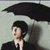 A person in a dark suit and white shirt holds a black umbrella against a light gray background, viewed from a side angle with a slightly pixelated appearance.