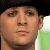 A pixelated close-up of a person's face with a black cap, light skin, and neutral expression against a blurred green and white background.