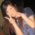 A pixelated image of a person with long brown hair wearing a blue top, making a hand gesture near their face against a dark background.