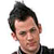 Static pixelated image of a person's head and shoulders with dark hair, visible facial features including eyes and nose, and partial clothing in black and brown tones against a white background.