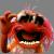 A pixelated red and orange creature with large eyes and an open mouth showing teeth, set against a blurred background.
