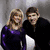 Two people pose together in a dark room with one wearing purple and the other black, while another person appears in red attire.