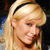 Pixelated close-up of a person with long blonde hair tied back by a black headband, wearing a dark top against a blurred background.