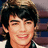 Pixelated close-up of a person with dark hair and light skin smiling, wearing a dark shirt with a light collar, against a blurred background with brown and blue tones.