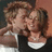 Two figures in close physical proximity with one figure's mouth near the other's cheek, both wearing white-collared dark shirts against a gradient background.