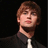 Pixelated portrait of a person with light brown hair, wearing dark clothing and a white tie against black background.