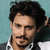 A person with dark curly hair and a mustache wears a white collared shirt under a gray jacket against a teal background.