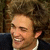 A person with curly brown hair wearing a dark suit jacket and white shirt, smiling broadly in four showing slight head movement