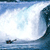 Static pixelated image of a large ocean wave curling over with white foam, featuring small dark shapes resembling boats or surfers in the lower-left corner against a blue sky background.
