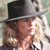 A pixelated close-up of a person wearing a dark wide-brimmed hat, with light brown curly hair visible beneath the brim, set against a blurred background.