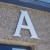 A large light blue capital letter A with dark blue and gray shading on a textured gray background with horizontal brown and blue bands.