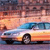 Pixelated image of a light-colored sedan with blue accents driving on a road under a sunset sky with orange and pink hues.