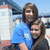 Two people in a blue and black shirt pose together against a blurred background with white structures.