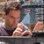A pixelated image shows a person with brown hair wearing a dark shirt, holding a light blue triangular object near their face against a blurred background.