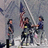 Pixelated static image of three figures in dark clothing with one figure raising an arm, set against a muted gray background.