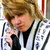 A pixelated image of a person with light brown hair holding a microphone, wearing a white jacket with black stripes over a black shirt.