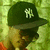 A person wearing a dark green cap with white NY logo, light brown hair, and red clothing against a green patterned background.