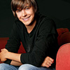 A person with light brown hair sits on a red couch, wearing a black long-sleeve shirt and light blue jeans, smiling with arms crossed.