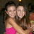 Two women in a close embrace, one wearing a bright pink sleeveless top against a dark background with blurred figures and warm lighting.