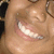 A person with light brown skin and glasses smiles broadly, showing white teeth and pink lips, with a dark background and a light gray area on the right.
