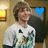 A person in a white graphic t-shirt stands against a warm-toned background with wall sconces, displaying a blue heart icon above their head.