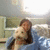 A person in a blue jacket holds a small light-colored dog against a blurred indoor background with green and white elements.