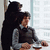 Two figures in a pixelated image: one standing behind another who sits at a table with hands clasped, both facing right.