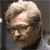 Pixelated close-up of a person with light-colored hair and glasses, wearing a dark shirt against a neutral background.