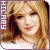 Pixelated static image of a smiling person with long blonde hair and dark brown side-swept bangs, featuring vertical text on the left edge against a purple border.