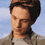 A pixelated image of a person with dark hair and light skin, wearing a gray jacket over a white shirt against a blue sky background.
