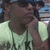 A person in a light green shirt with dark text holds their hand near their face, wearing sunglasses and a cap.
