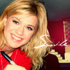 A person with shoulder-length blonde hair wearing a red top, holding a pen-like object, with a red wall and decorative background elements visible.