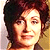 Pixelated portrait of a person with dark curly hair, light skin tone, and neutral expression against gradient background.