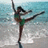 A woman in a green swimsuit performs a high kick on a beach with ocean waves in the background.