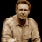 Pixelated static image of a person with light brown hair wearing a beige zip-up jacket over a dark shirt against black background.