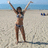A woman in a bikini stands on a beach with arms raised, facing the camera against a blue sky and ocean backdrop.
