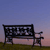 Pixelated image of a dark metal bench with ornate backrest against a gradient sky transitioning from blue to pink, set on a grassy area.