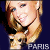 Pixelated image of a smiling woman with blonde hair and blue eyes, wearing a dark top against a solid dark background.
