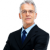 Pixelated image of a person in a dark suit with a light blue shirt and dark blue tie against a gray background with scattered white pixels.