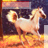 Pixelated image of a light-colored horse standing on a yellow-orange ground with dark background, head turned right, ears forward, tail visible.
