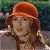 A pixelated figure with reddish-brown hair wears an orange cap and a brown top against a blurred green and dark background.