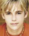 Static close-up portrait of a person with light brown hair, wearing an orange shirt and beaded necklace against a blurred background.