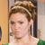 A pixelated close-up of a person with light brown hair styled in an updo, wearing a white top with green accents, against a blurred indoor background.