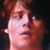 Pixelated close-up of a person's face with reddish-brown hair, light skin, and a neutral expression against a dark background.